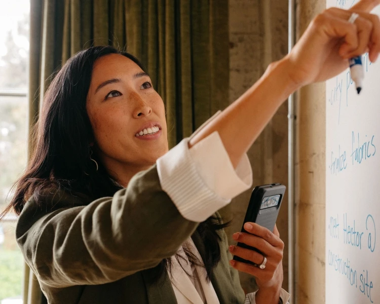 Woman writing on whiteboard in an office setting. She is holding a smart phone and a marker. The window is providing natural light.