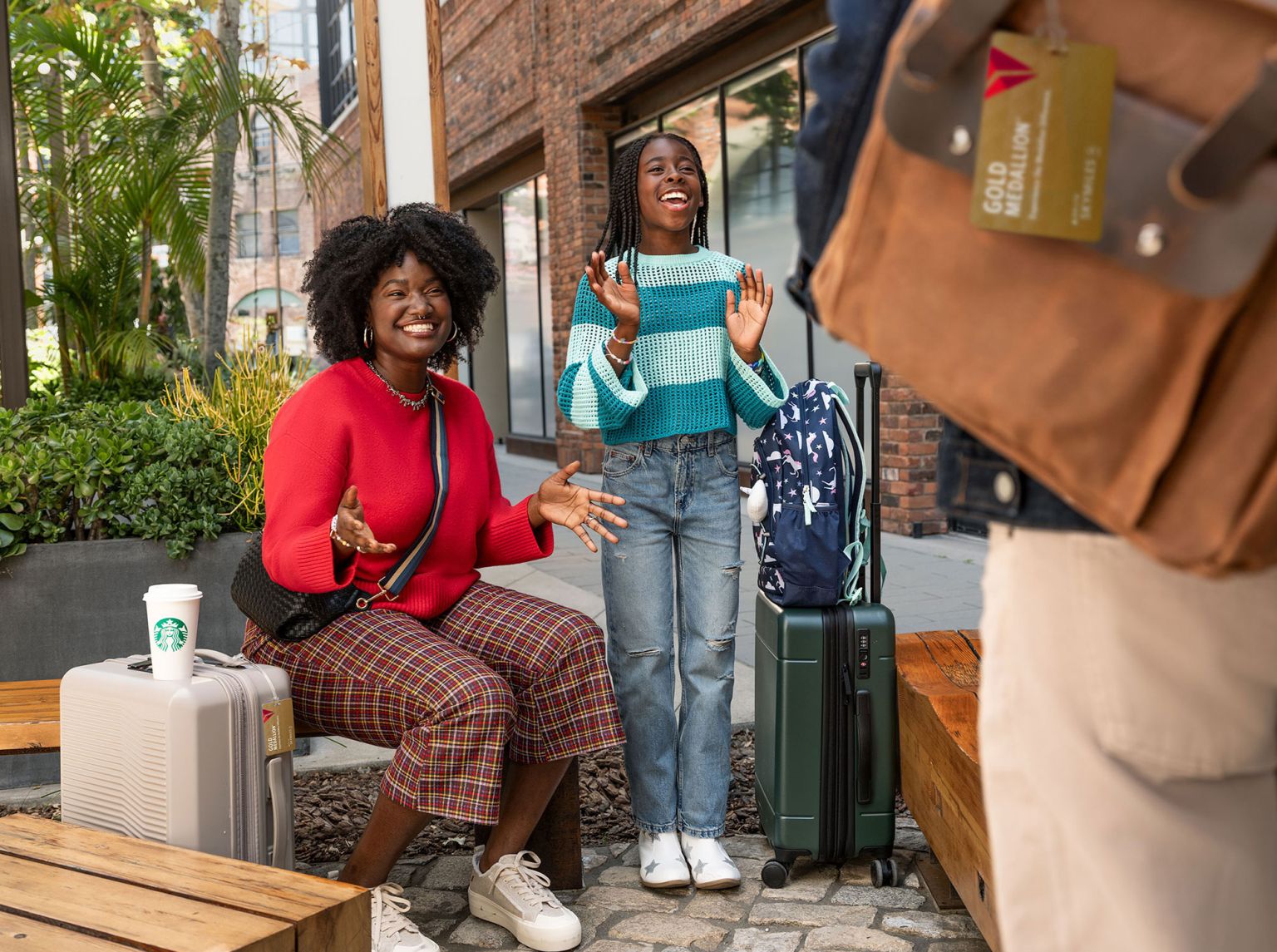 A happy mother and daughter sit next to luggage while a third person stands nearby in an outdoor, brick-paved setting with greenery.