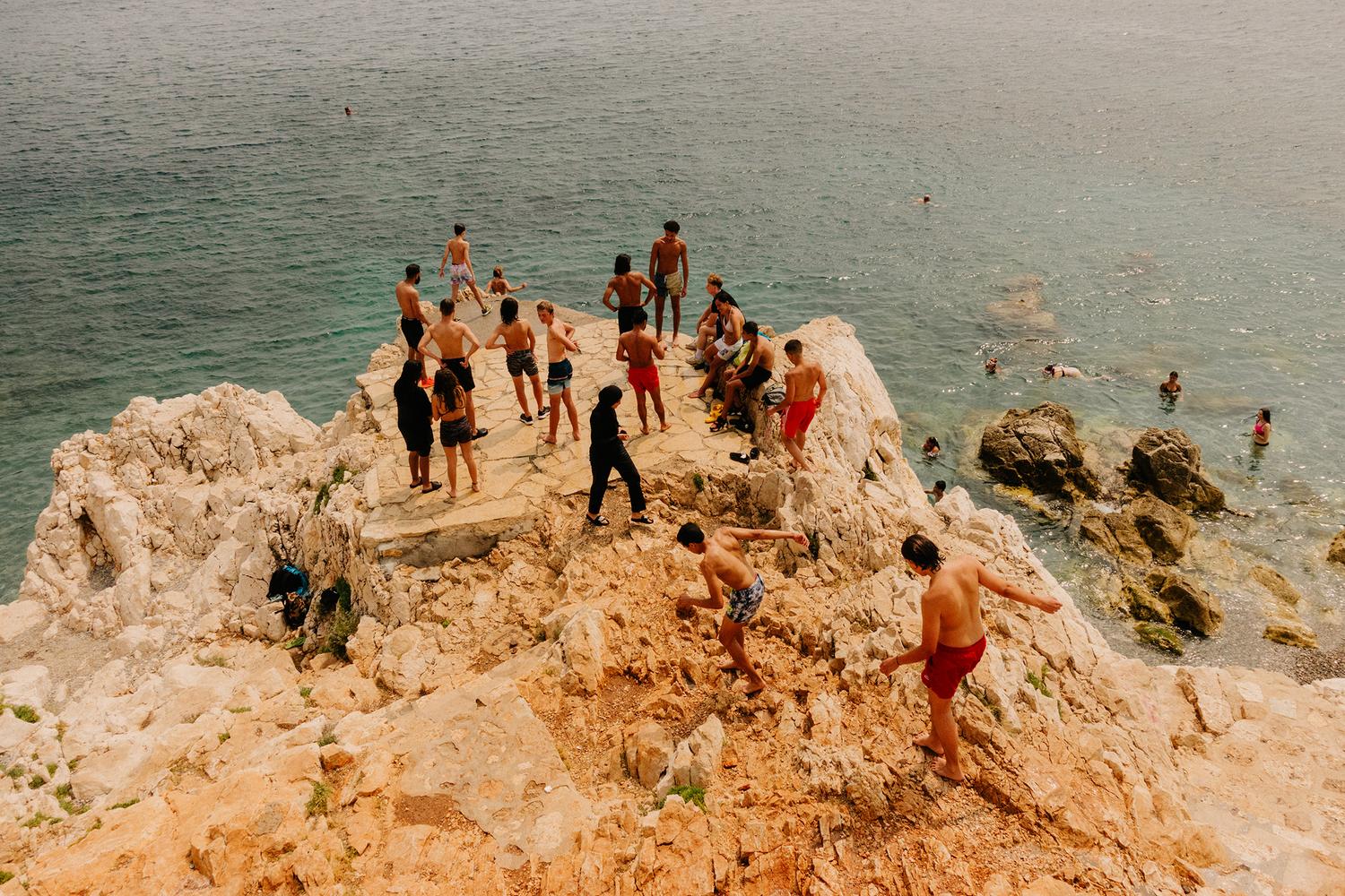 A group of people of different ages gather on a rocky cliff overlooking the ocean, swimming, standing, and jumping into the water.