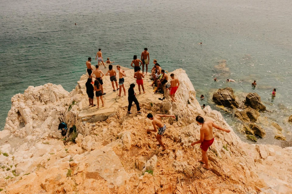 A group of people of different ages gather on a rocky cliff overlooking the ocean, swimming, standing, and jumping into the water.