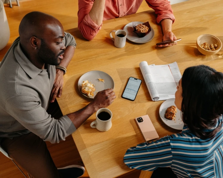 Overhead shot of three people at a table with food, drinks, phones, and papers, creating a casual, productive, and social atmosphere.