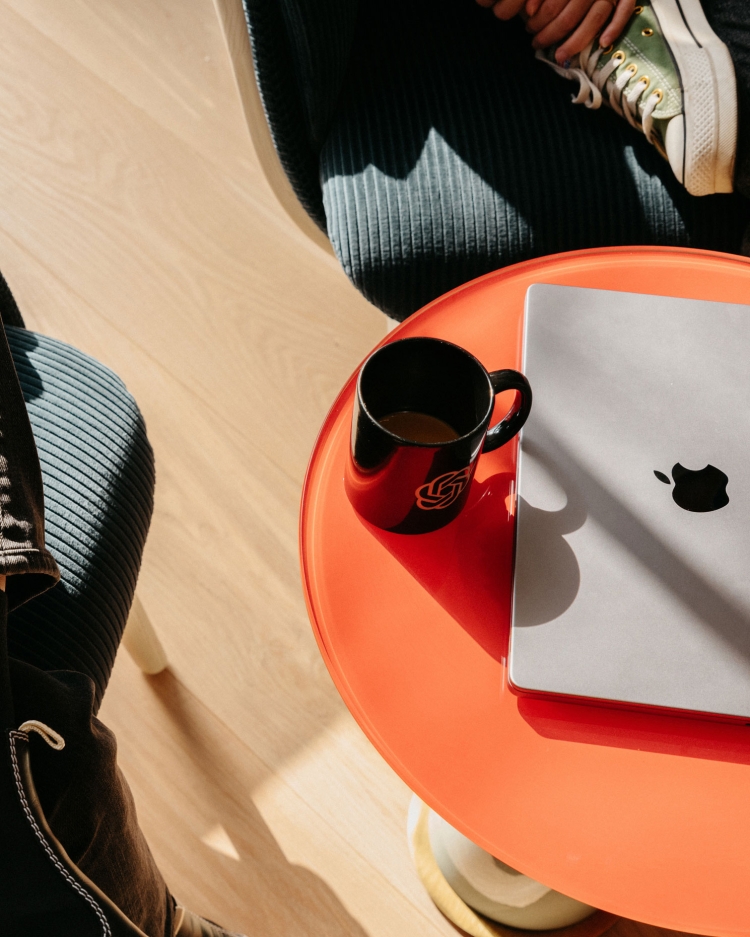 Laptop and coffee mug on a round orange table next to two chairs indoors in a brightly lit scene, capturing a casual workspace.