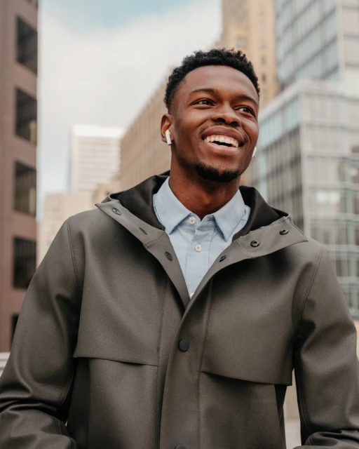 Smiling Black man in gray hooded jacket stands in urban setting with modern buildings, wearing wireless earbuds and looking upwards.