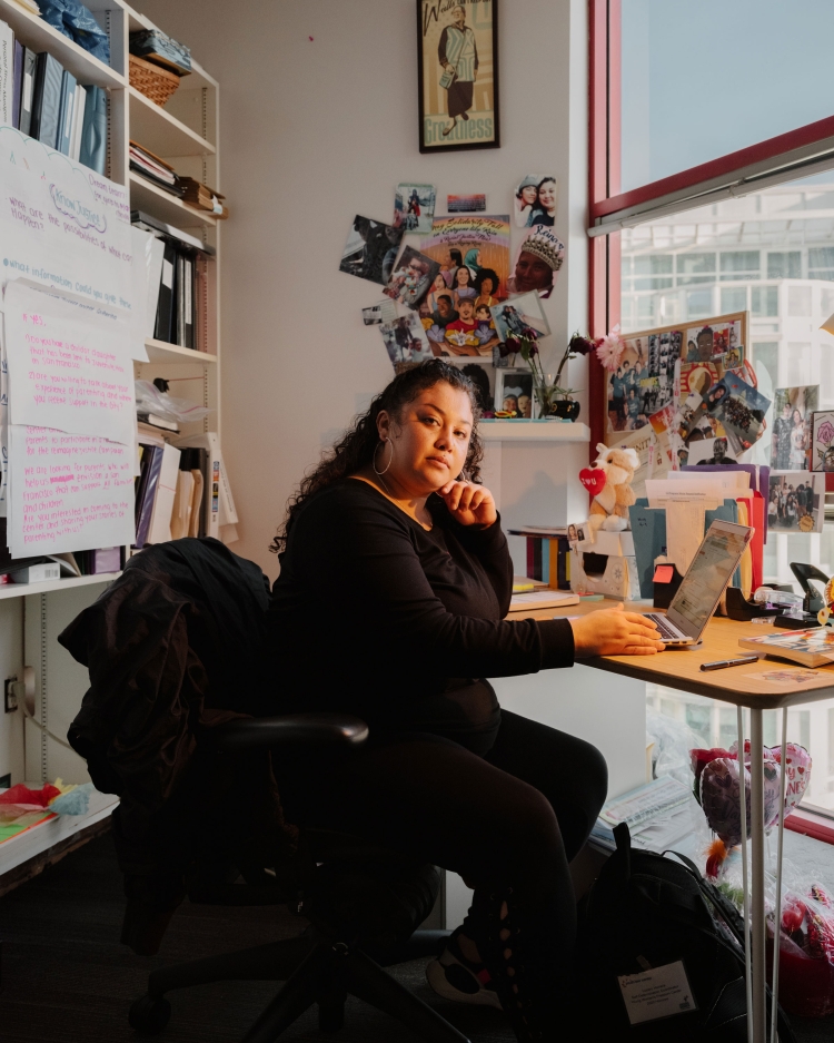 Adult woman in black outfit sits at a desk indoors, working on a laptop, with family photos and a bookshelf in the background.