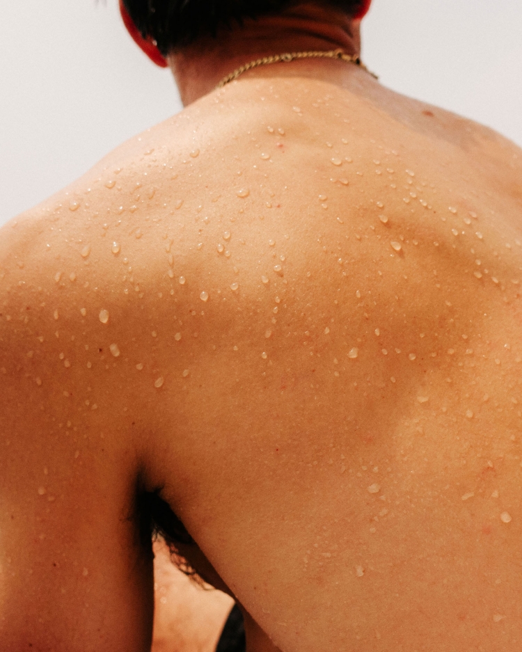 Close-up of a man's bare back with water droplets, wearing a gold chain. Against a plain white background.