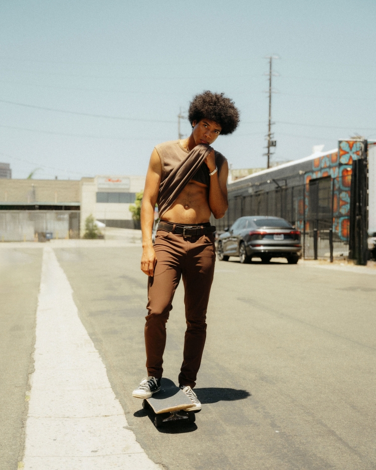 Young adult male poses on a skateboard on an asphalt street, wearing brown pants and a sleeveless brown shirt on a sunny day.