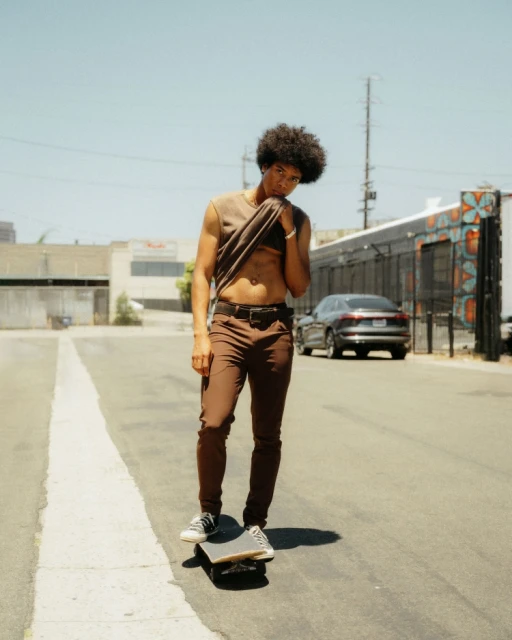 Young adult male poses on a skateboard on an asphalt street, wearing brown pants and a sleeveless brown shirt on a sunny day.