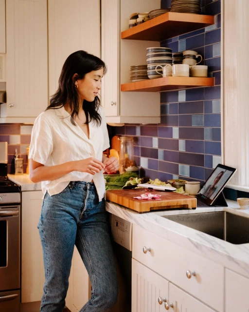 A young Asian woman stands in her bright kitchen, preparing food while video-chatting on a tablet. Radishes and green vegetables are on a cutting board.