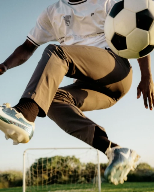 Action shot of a soccer player in mid-air kicking a soccer ball against a bright blue sky with a soccer net and green trees in the distance.