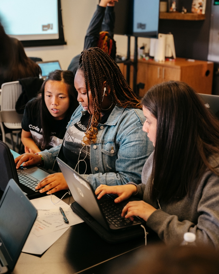 Three diverse women collaborate using laptops at a table, likely working on a project or studying together in an indoor setting.