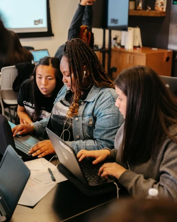 Three diverse women collaborate using laptops at a table, likely working on a project or studying together in an indoor setting.