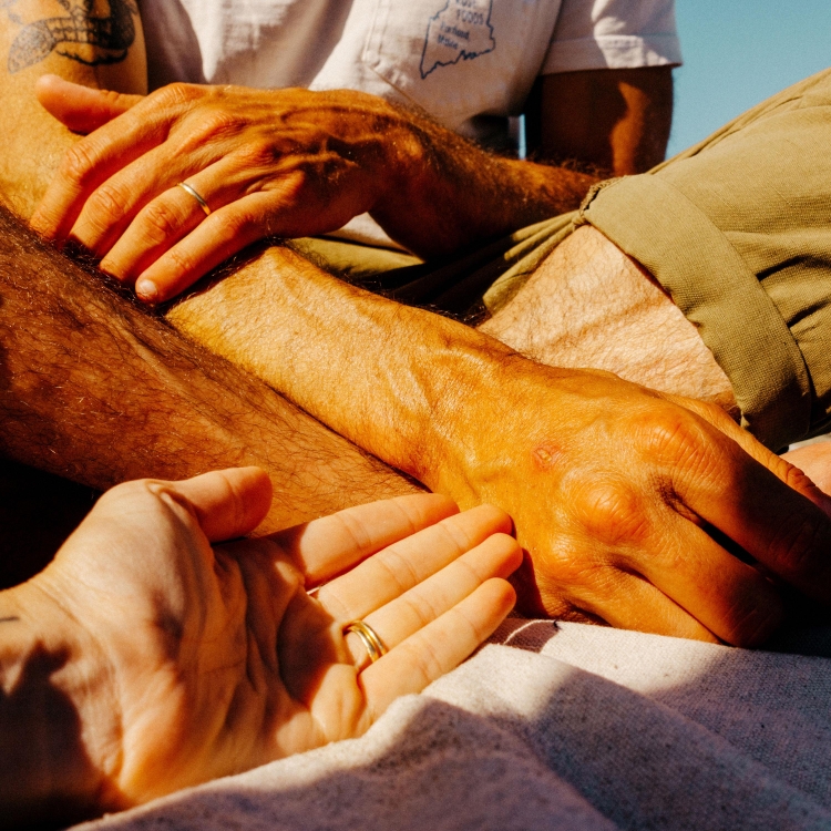 Close-up shot featuring two people's hands, one resting palm-up in the other's palm, highlighting wedding rings and sunlit skin tones.