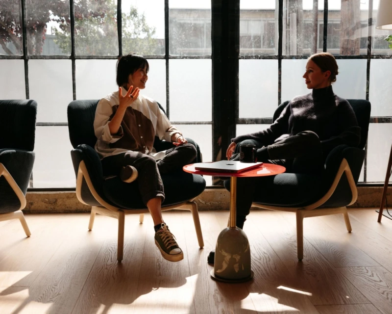 Two women are sitting in modern chairs having a conversation near a window, with a small table between them holding a laptop.