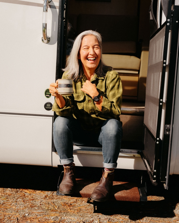 Smiling older woman with grey hair sits on the steps of a camper with a coffee mug outdoors, wearing a green plaid shirt and jeans.
