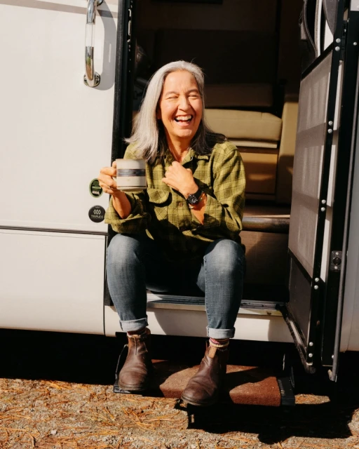 Smiling older woman with grey hair sits on the steps of a camper with a coffee mug outdoors, wearing a green plaid shirt and jeans.