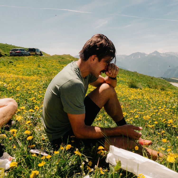 Adult male sitting in a field of yellow flowers on a sunny day, with a mountain range and cars visible in the background.
