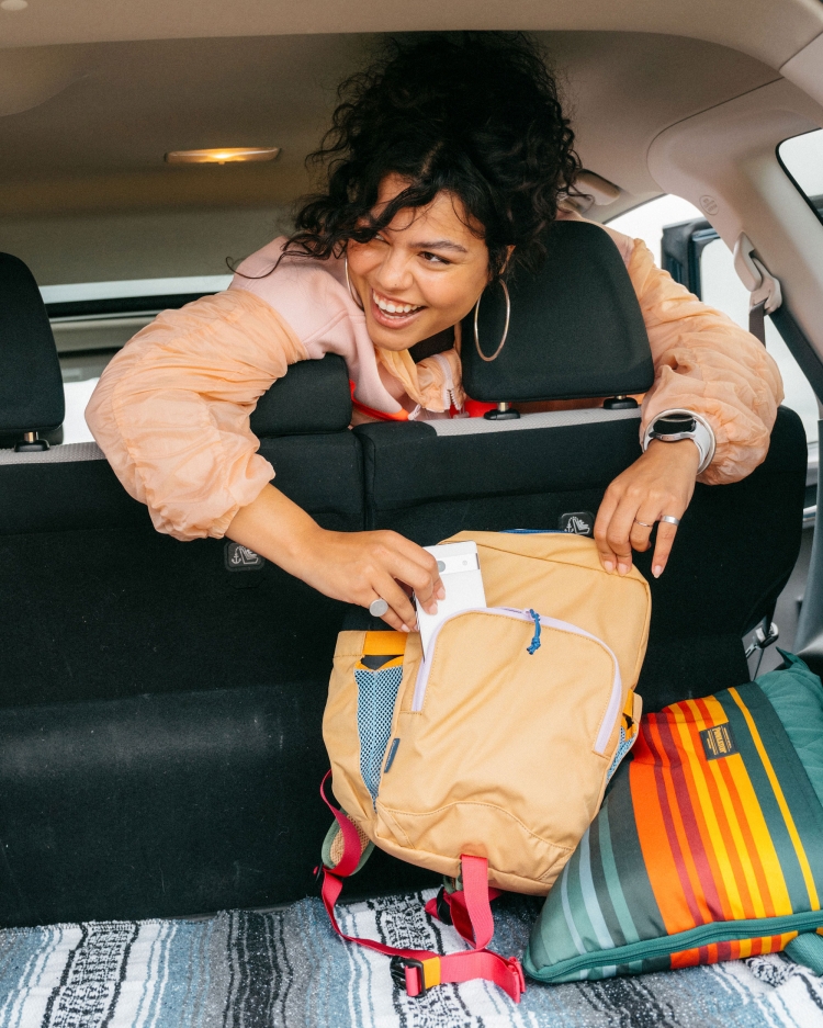 Smiling woman in car, taking tissue out of backpack. Casual peach-color jacket, patterned blanket, and car interior.