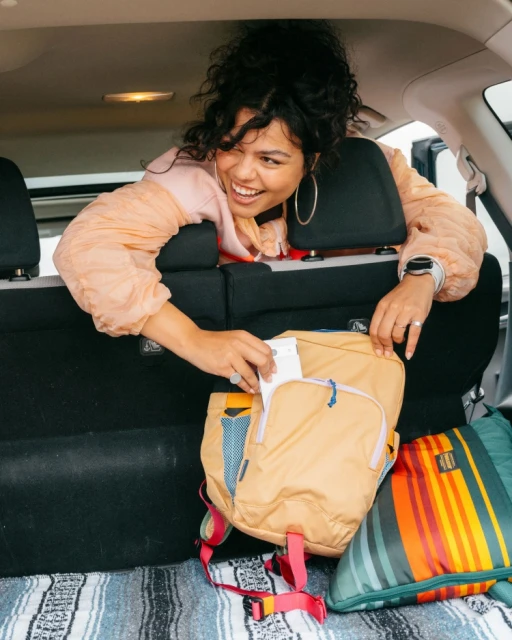 Smiling woman in car, taking tissue out of backpack. Casual peach-color jacket, patterned blanket, and car interior.