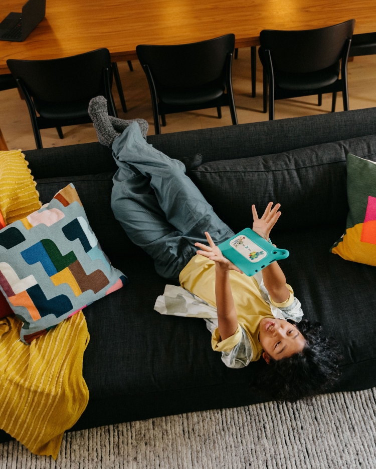 A young child with dark hair is laying on a dark gray couch and looking at a teal-colored tablet, with colorful decorative pillows nearby.