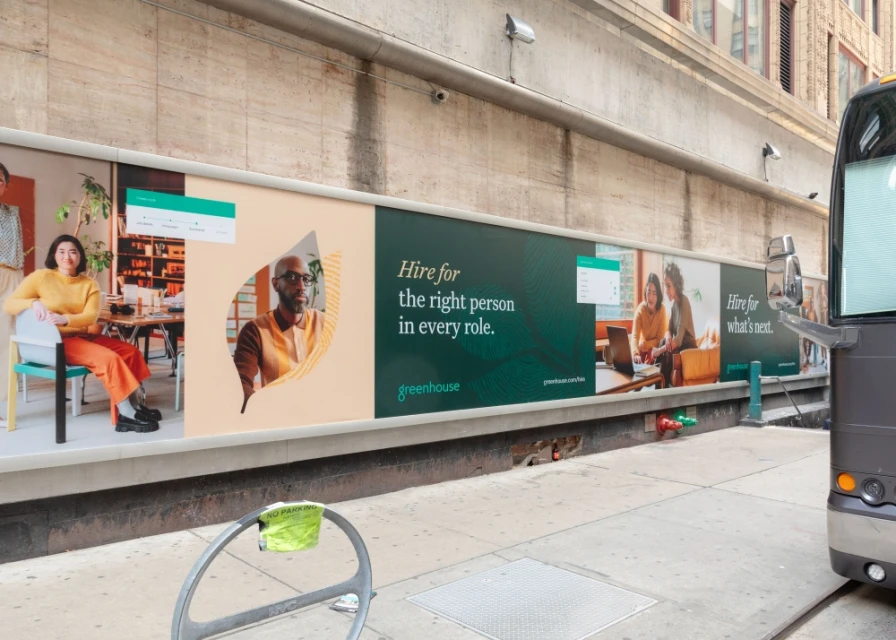 Outdoor billboard ad with images of people, text about hiring, and the Greenhouse logo, next to a city street and bus on the right.