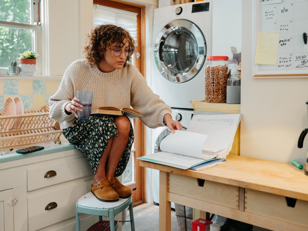 Woman with curly hair sits on stool in kitchen reading book, holding glass, with washing machine and home decor in background.