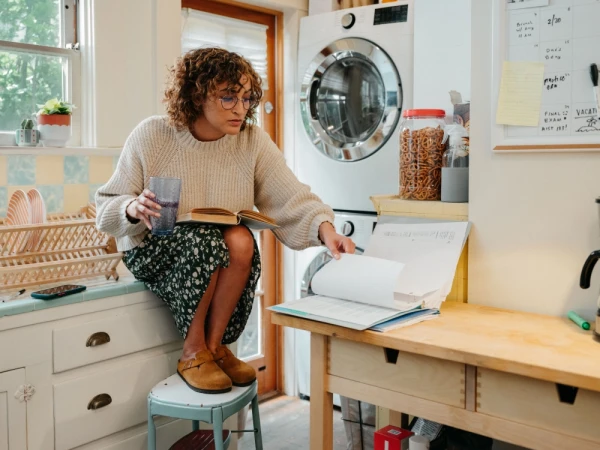 Woman with curly hair sits on stool in kitchen reading book, holding glass, with washing machine and home decor in background.