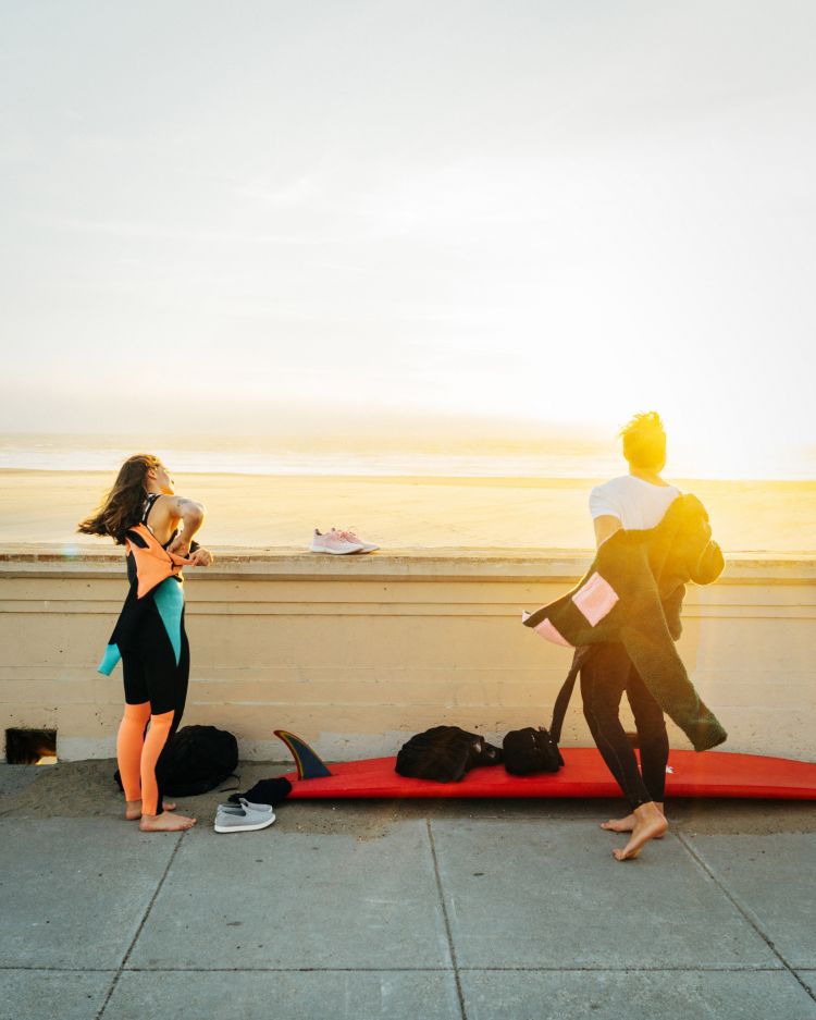 Two surfers in wetsuits prepare near the beach; one taking off a wetsuit, surfboard at her feet on a sunny day near the ocean.