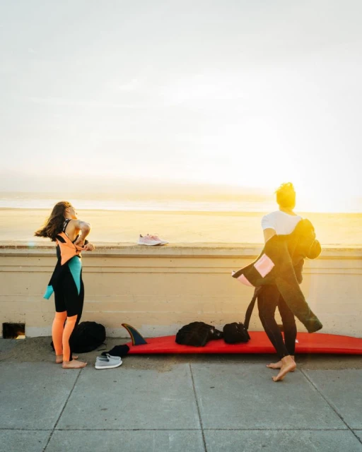 Two surfers in wetsuits prepare near the beach; one taking off a wetsuit, surfboard at her feet on a sunny day near the ocean.
