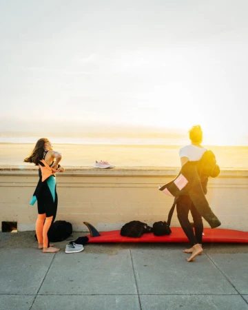 Two surfers in wetsuits prepare near the beach; one taking off a wetsuit, surfboard at her feet on a sunny day near the ocean.