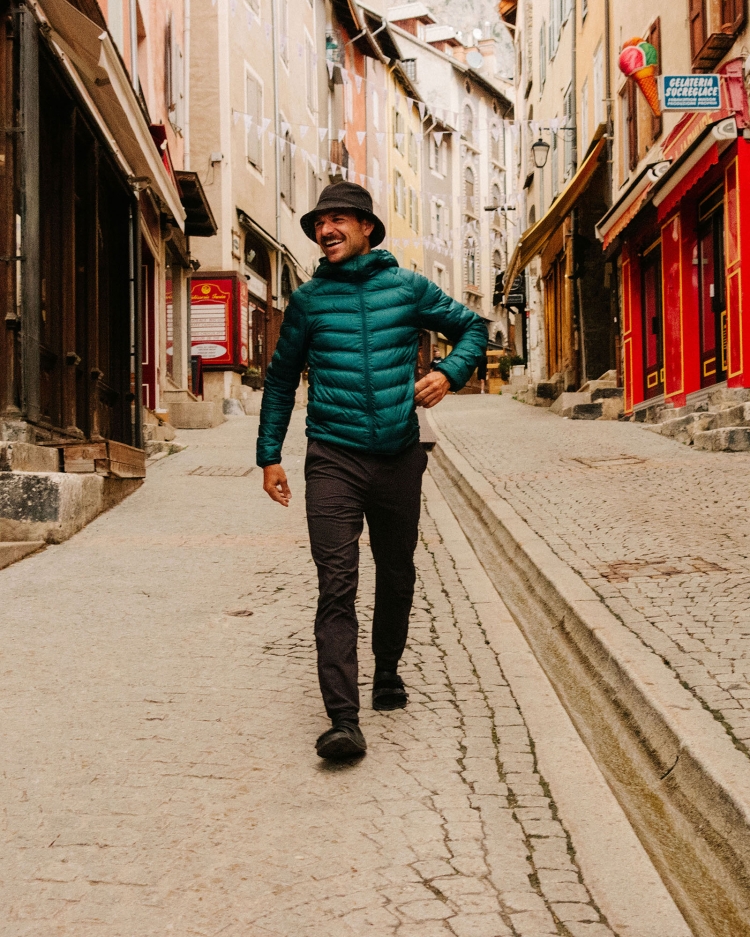 Adult male in green puffer jacket walks down cobblestone street lined with buildings on a sunny day with blue sky and clear light.