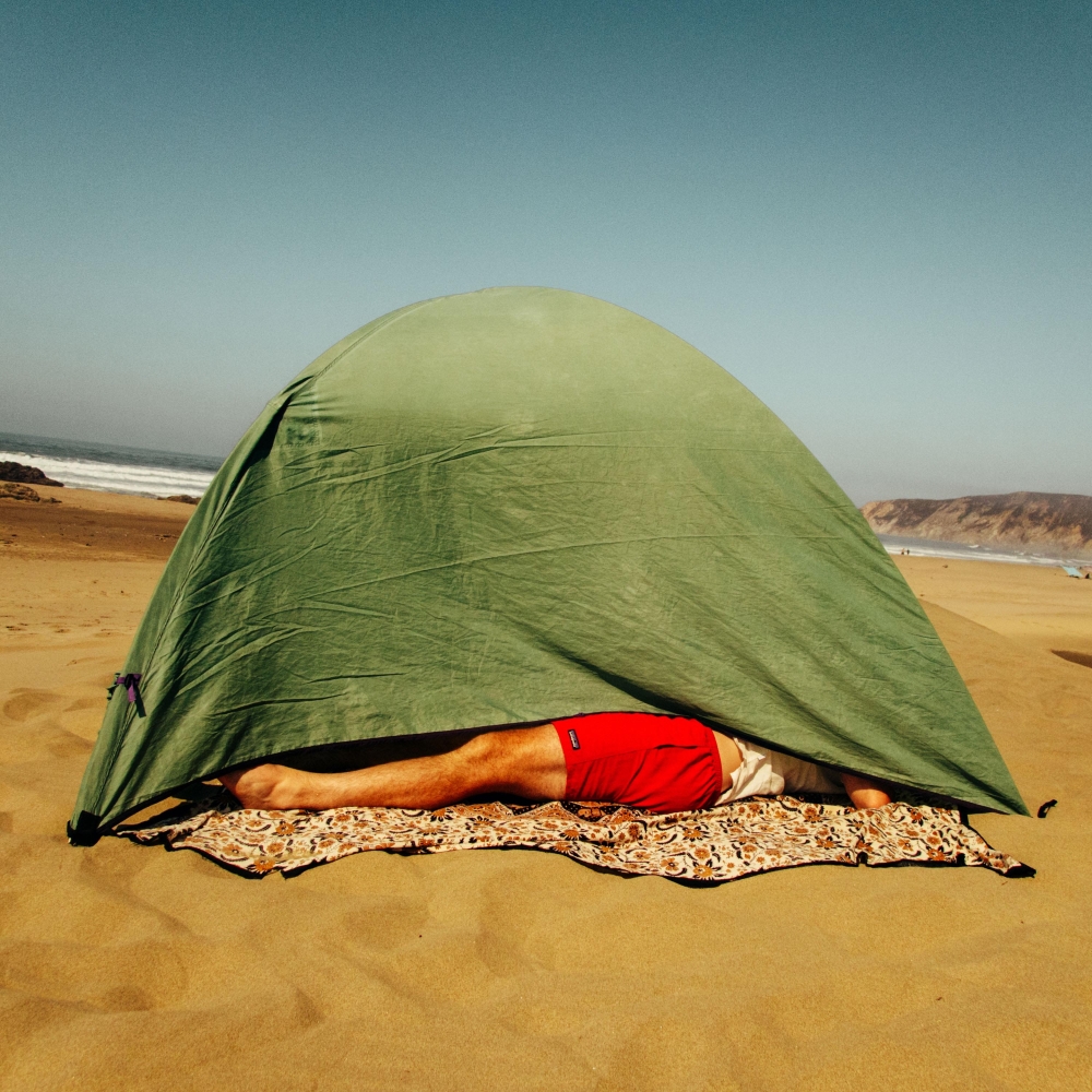 Man in red shorts lying on a blanket under a green tent on a sandy beach with waves and coastline in the background on a sunny day.