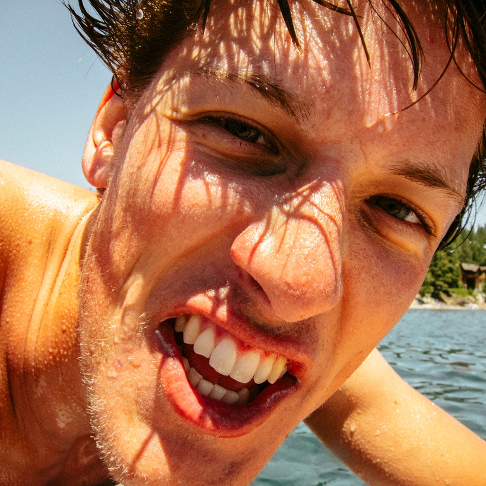 Close-up of a grinning adult male with wet hair, captured outdoors on a sunny day, with a water body and trees in the background.