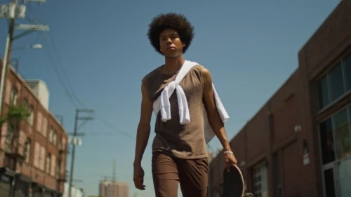 An African-American man walks on a street, wearing a brown tank top and brown shorts. He carries a skateboard, with city buildings in the background.