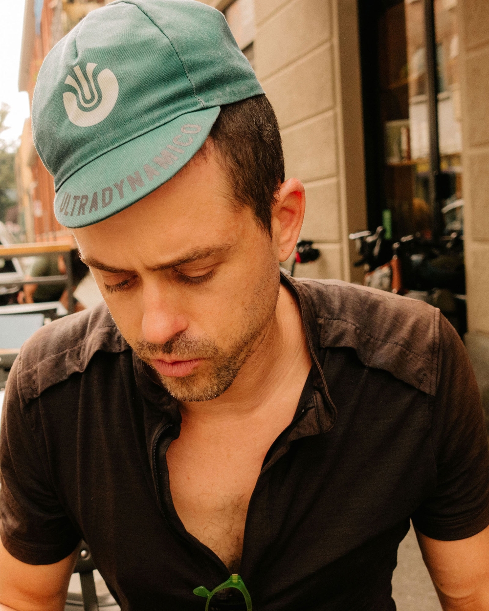 Man wearing a teal cycling cap with a logo, seated outdoors at a cafe or restaurant, captured in a medium shot during daylight hours.