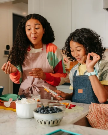 Two young people in aprons preparing food at a kitchen counter with bowls of blueberries and other ingredients, illuminated by bright light.