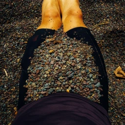 Close-up perspective of a person sitting outdoors with legs covered in dark pebbles and stones, wearing dark shorts.