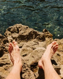 Bare feet resting on a rocky coastal surface above dark shimmering water by Jake Stangel