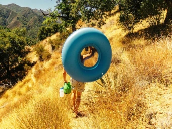 A person walking up a dry grassy hill carrying a large blue inner tube and a green pouch.