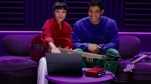 Two musicians, an adult male and an adult female, sit on a purple couch with guitars in a music studio with purple lighting.
