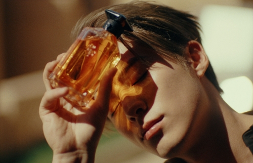An adult man holds a perfume bottle in front of his face, with the bottle reflecting light and creating shadows on his face in warm tones.
