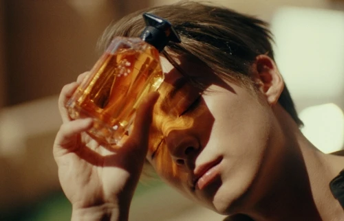 An adult man holds a perfume bottle in front of his face, with the bottle reflecting light and creating shadows on his face in warm tones.