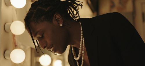 Adult male poses indoors in a black jacket and pearl necklace, lit by vanity lights for a fashion or celebrity and entertainment context.