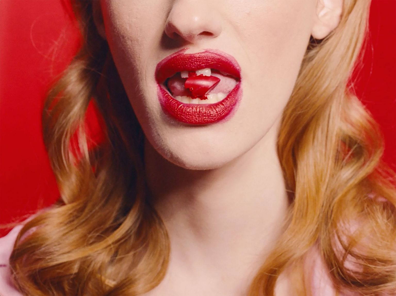Close-up of a fair-skinned adult female face with bright red lipstick, holding a red candy in her mouth, set against a vivid red backdrop.