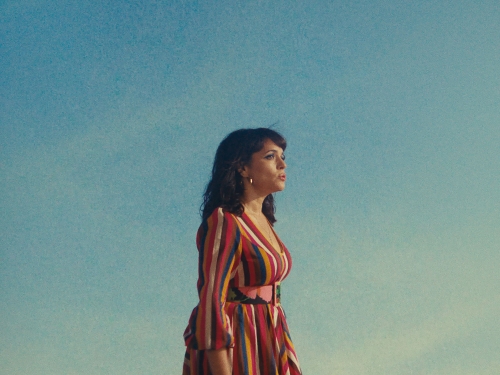 Adult woman in colorful dress poses outdoors in a medium shot against a plain blue sky during the day in natural light.