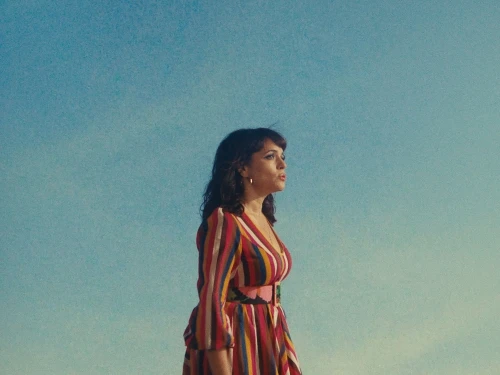 Adult woman in colorful dress poses outdoors in a medium shot against a plain blue sky during the day in natural light.