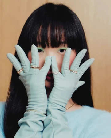 Close-up portrait of a woman with long dark hair wearing sage green leather gloves up to her forearms and rings on her fingers.