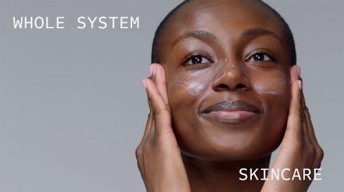 Adult female with dark skin applies skincare cream to her face, smiling softly against a neutral gray background, promoting beauty and wellness.