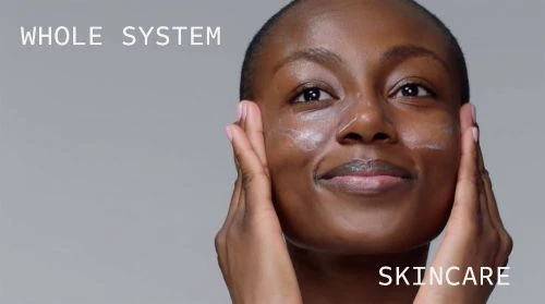 Adult female with dark skin applies skincare cream to her face, smiling softly against a neutral gray background, promoting beauty and wellness.