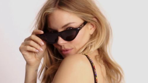 Blonde woman poses in a close-up studio shot, wearing black sunglasses and a colorful beaded top against a bright white background.