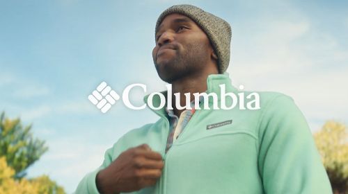 A man in a beanie and fleece jacket stands outside under a blue sky, with the Columbia logo in the foreground. The focus is on the apparel.